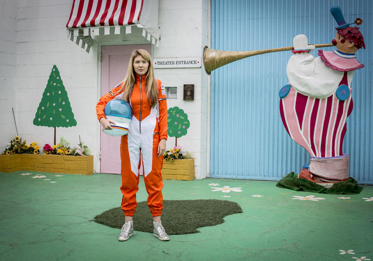 Beatie Wolfe wearing an orange space suit, with a colourful house behind her