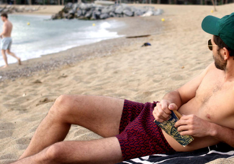 A man reclines on a beach in dark pink shorts and a teal cap, holding a book and looking at another man by the sea