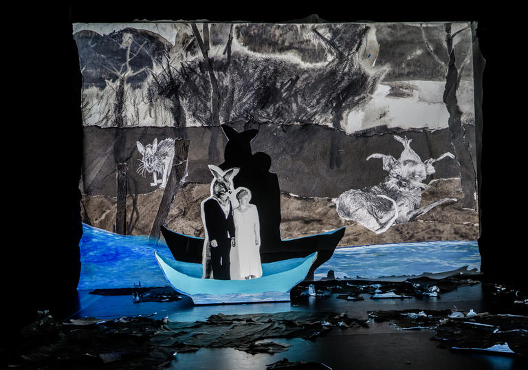 A figure that is half-man, half-kangaroo is standing in a boat with Fleur Elise Nobel's unnamed character. They look like figures in a pop-up book, against a paper painted backdrop and vivid blue colour in the water. Their shadows are prevalent against the paper backdrop.  