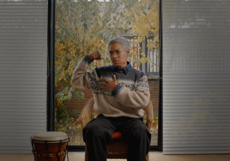 Glenn Copeland flexes a bicep in a grey and cream woolly jumper, in front of a window, next to a drum