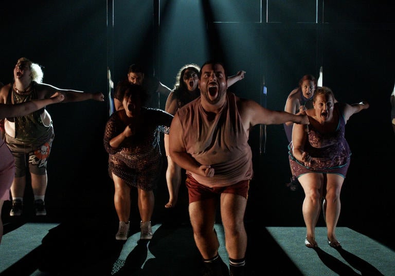 a dance troupe of dancers with larger bodies dances some choreography in a dark room 
