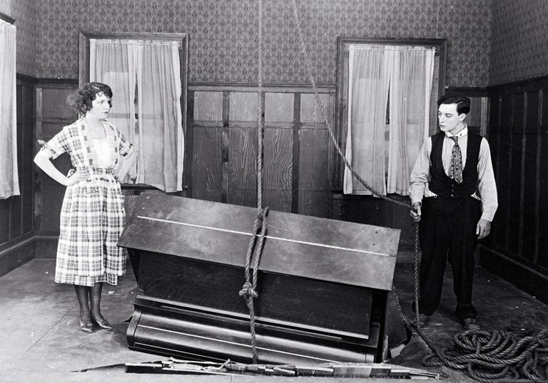 Buster Keaton and Sybil Seely stand in a room next to a piano which has crashed partially through the floor. 