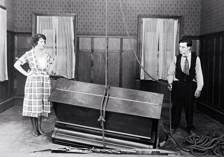Buster Keaton and Sybil Seely stand in a room next to a piano which has crashed partially through the floor.