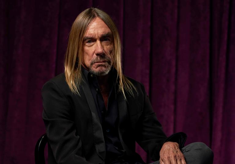 Iggy Pop, with long hair and a beard, is sitting on a chair in front of a velvet curtain