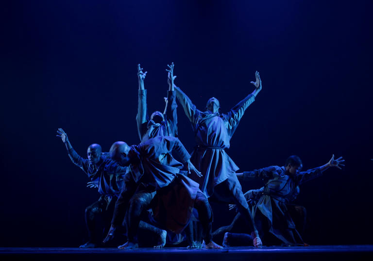 dancers in a clusters reach out to the sides/the ceiling with blue lighting 