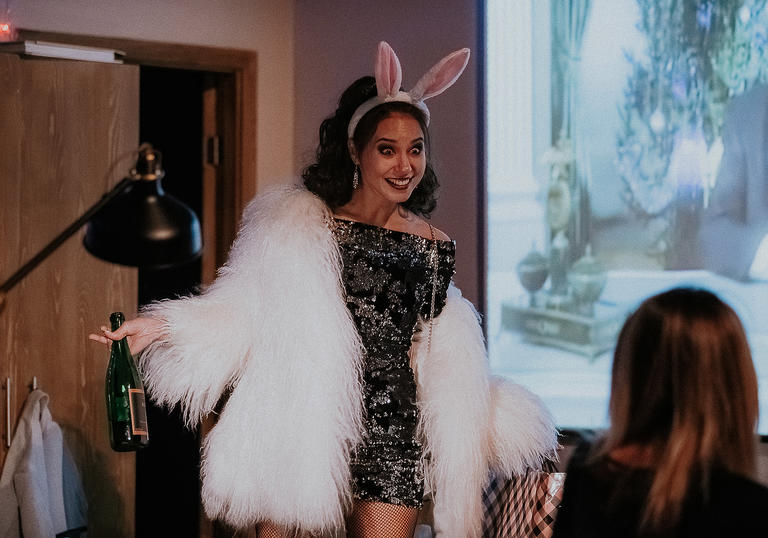 woman wearing bunny ears and a big white fur coat parades in front of her friend