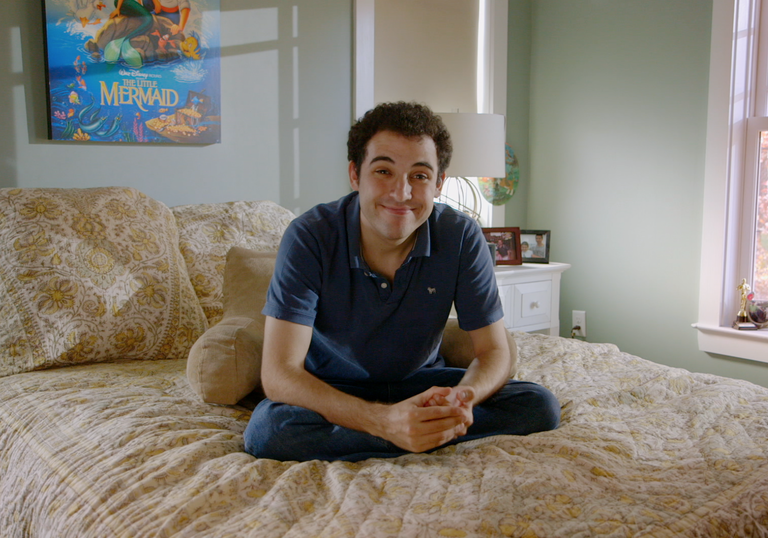 A young man sits on a bed smiling, with a poster of the Little Mermaid behind him