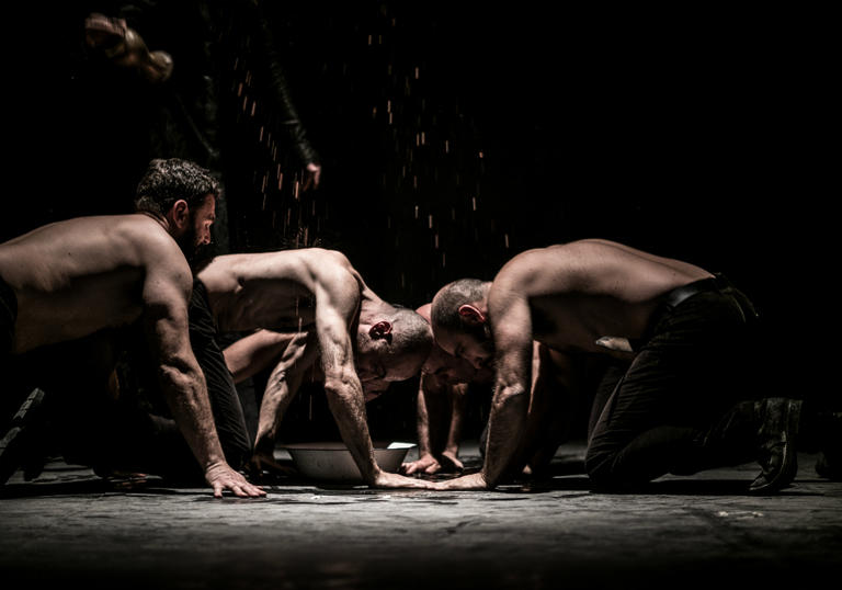 four topless men in a dark room appear to be eating from the floor