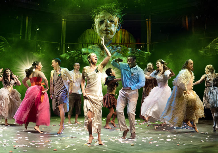 people dance in magical outfits with a giant green forest puppet behind them