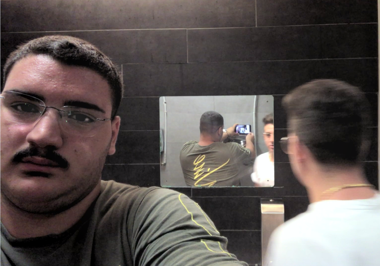 A man takes a selfie in a bathroom and we see this reflected in a mirror opposite, while a friend looks directly into the mirror