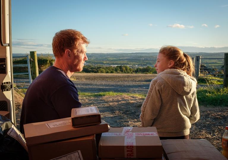 Ricky and his daughter sitting at the back of his van with the doors open looking out at the hills