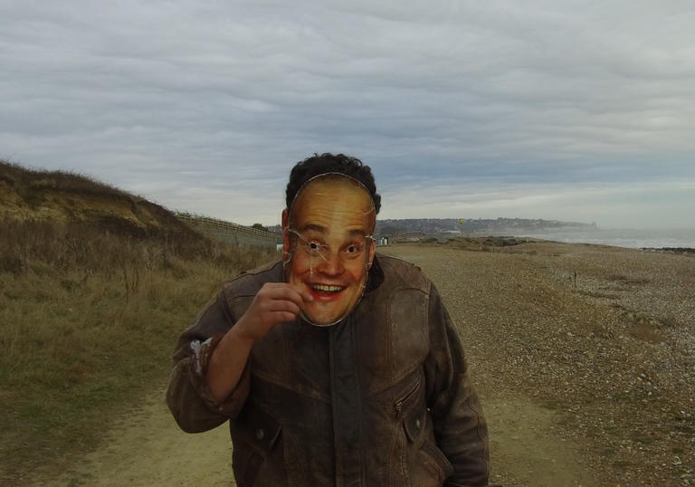 A man iin a green field holds a mask of a face over his own face