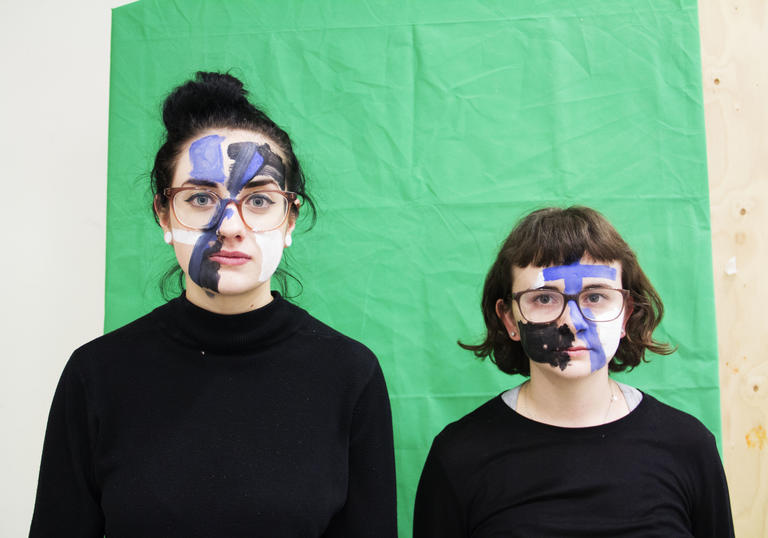 Two women, standing in front of a green screen with paint on their face