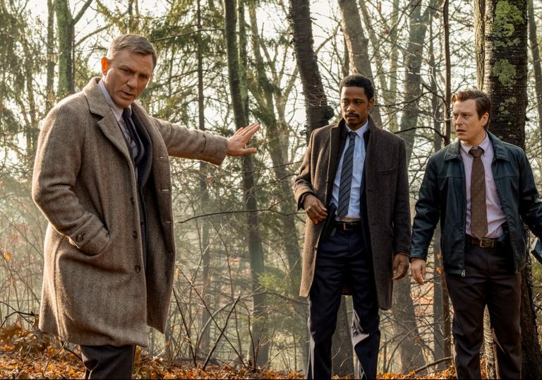 Daniel Craig in a cream overcoat stands in the woods with Lakeith Stanfield, in Rian Johnson's Knives Out