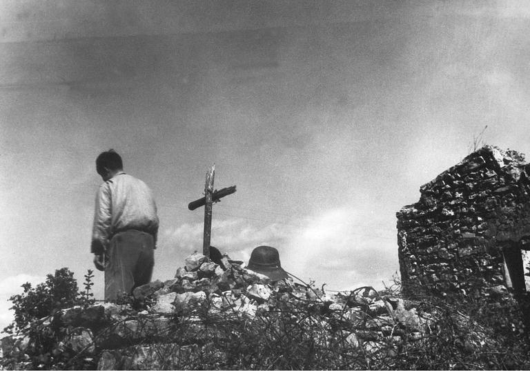 A black and white image of a boy with his back to the camera, by a makeshift grave