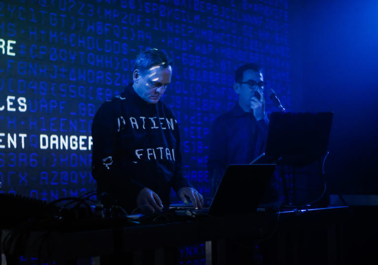 Alva Noto and Anne-James Chaton performing with a laptop and microphone. Words are projected behind them and onto their clothes