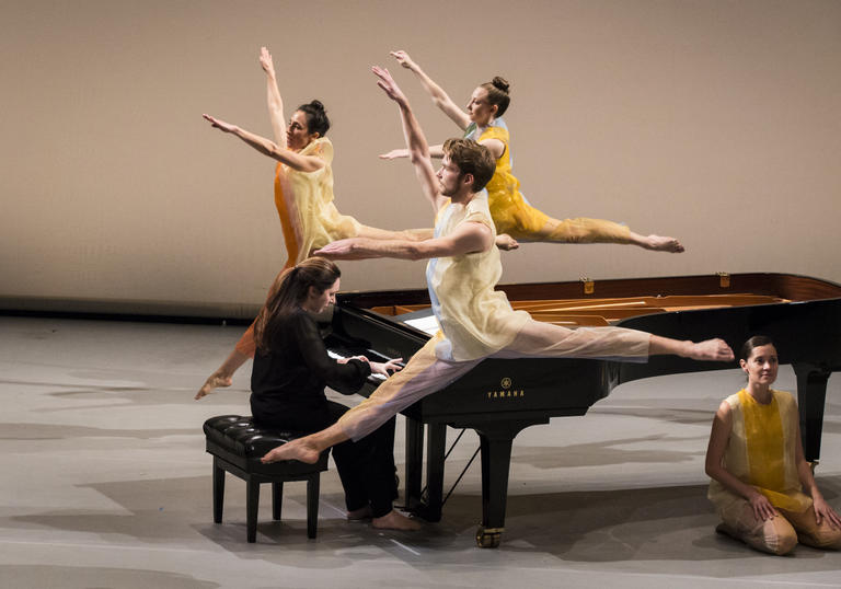 three dancers in yellow leap around a grand piano, one dancer is sat on the floor