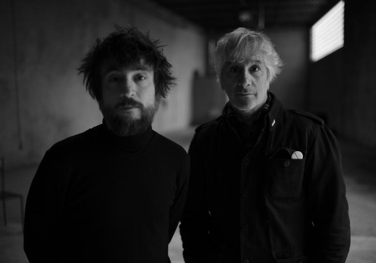 A black and white photo of Lee Ranaldo and Raul Refree in an empty concrete room, wearing black shirts