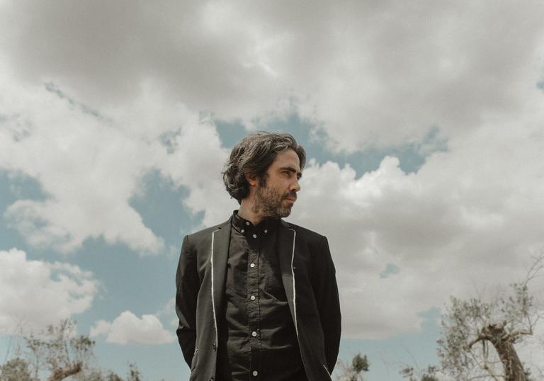 Patrick Watson wearing a black shirt and black jacket looking over his left shoulder, the sky behind him is cloudy