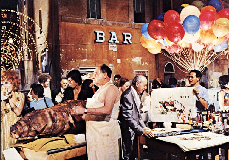 a market in a Rome square, there are balloons, meat and tables with people milling around