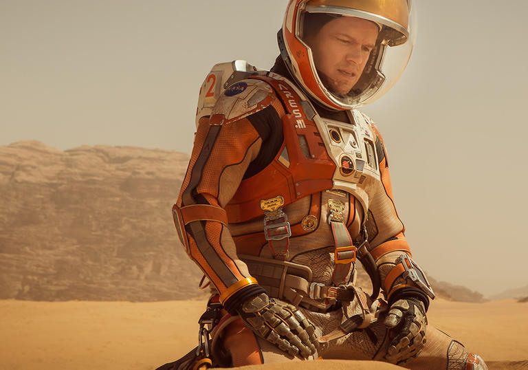 Matt Damon in an orange space suit on a dusty, desert-style landscape in The Martian