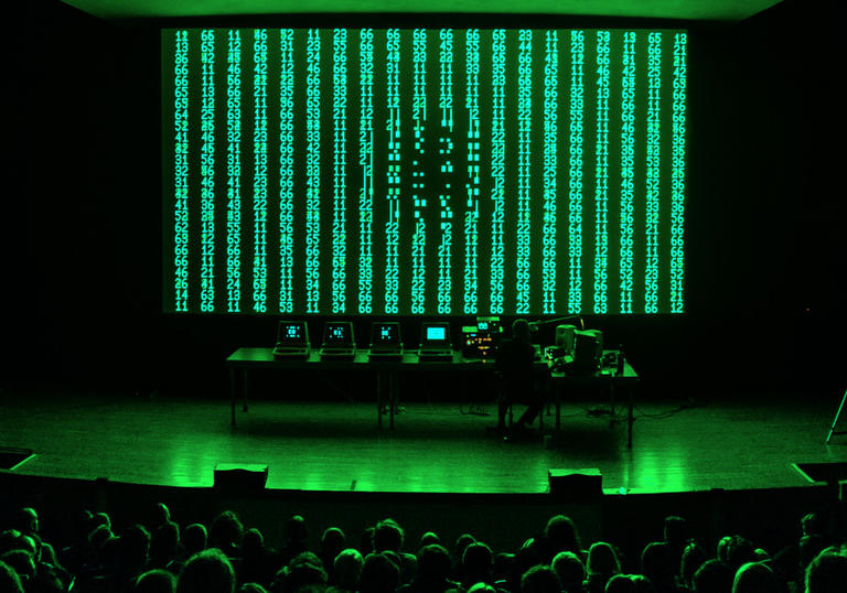 image of live performance shows robert henke standing in front of green and back digital screen with a bank of computers and electronics set up