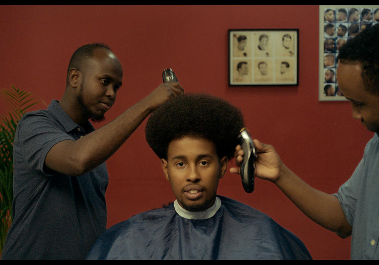 a man sitting in the barber chair with two barbers cutting his hair