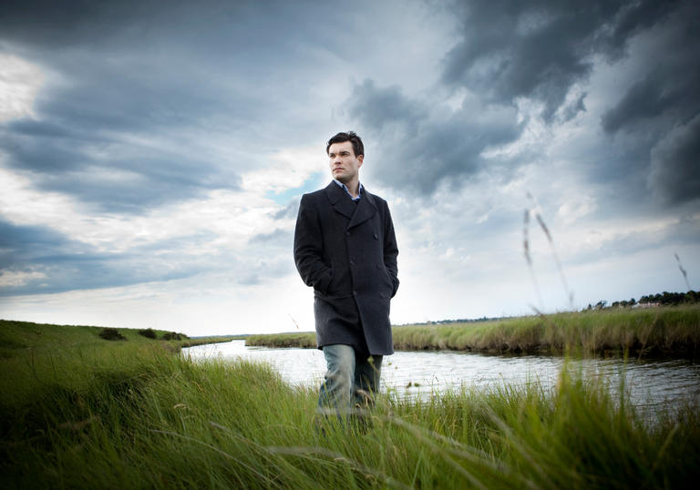 An image of Ryan standing on the grassy river bank, a dramatic grey sky overhead 