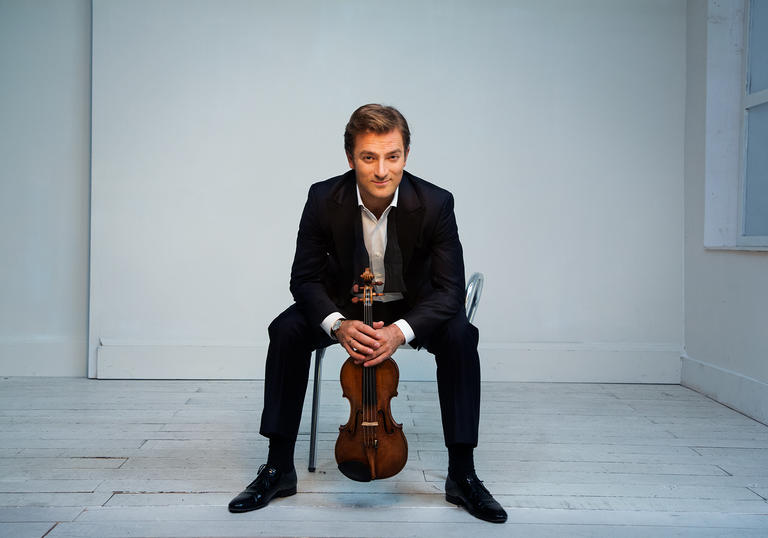 Renaud sitting on a chair in a white room with a cheeky grin on his face, clutching his violin