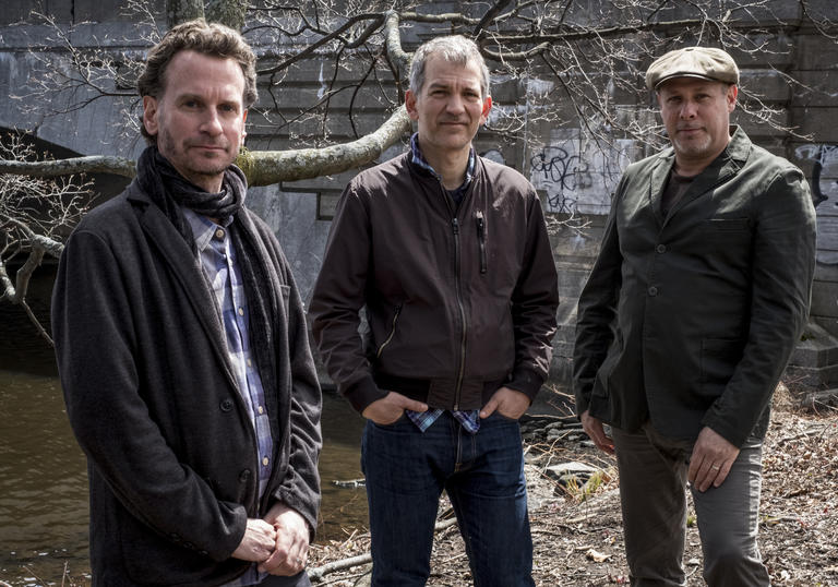 the trio stand next to a canal. They are dressed casually and it looks like autumn/winter