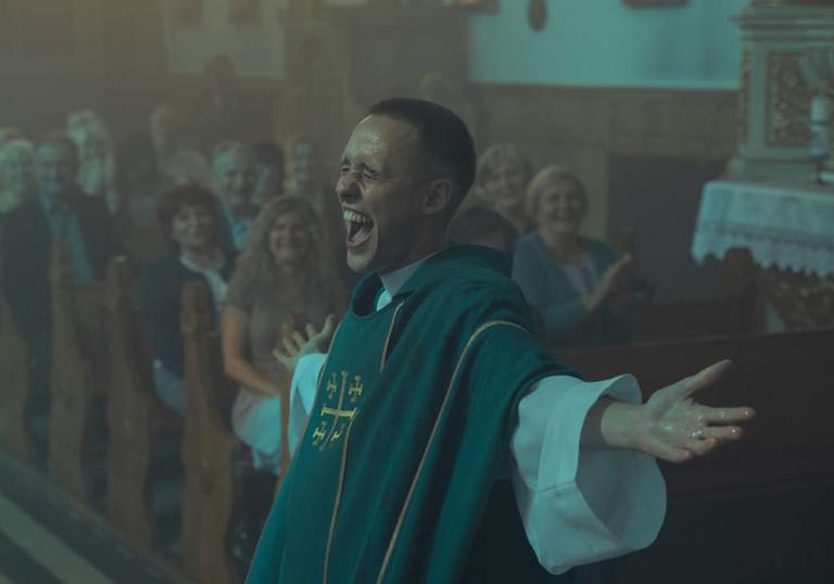 a man dressed as a priest stands in front of a congregation laughing