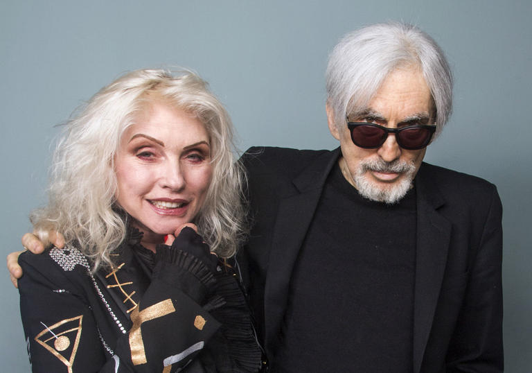 Debbie Harry and Chris Stein standing against a grey backdrop. Debbie is wearing an excellent suit, Chris is dressed in black and wears sunglasses. They have their arms around each other