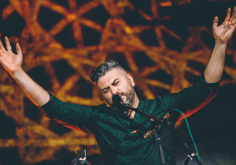 photo of Damir Imamovic performing live. He sings into the microphone and his arms are raised. The background is black and yellow, he wears a dark green shirt.
