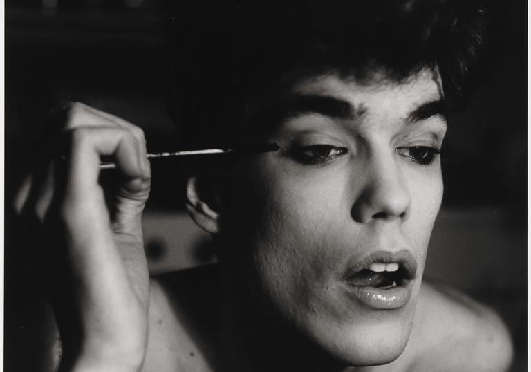 An black and white image of a man open mouthed, as he concentrates on putting on mascara