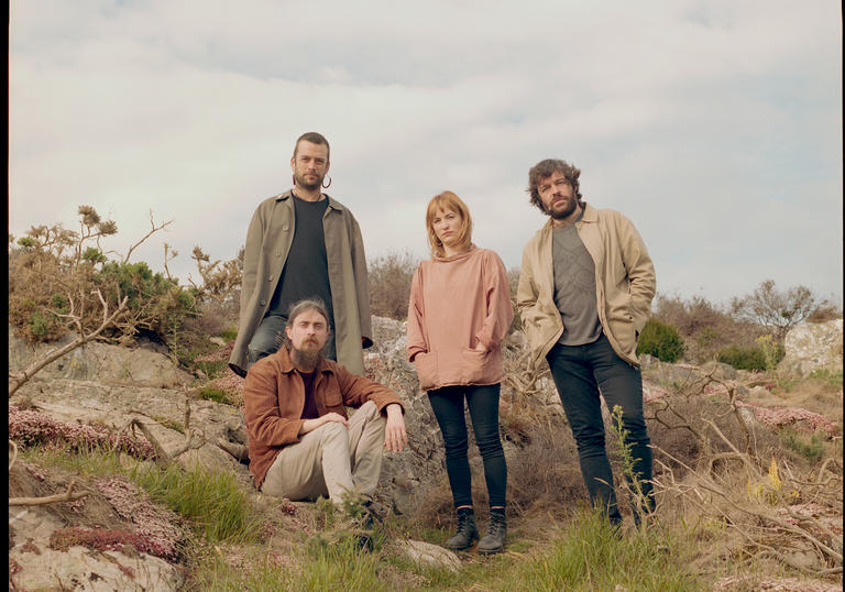 photo of the 4 band members in the countryside. They are surrounded by grass and rocks. 3 are standing, one is sitting and they face the camera