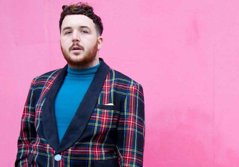 An image of Scotte standing against a vivid pink wall wearing a suave tartan two-piece suit