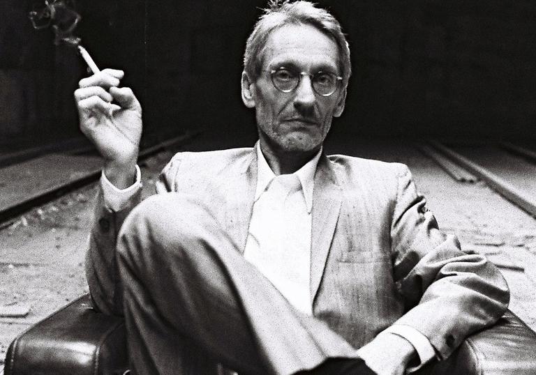 black and white photo of Manuel Göttsching sitting in a chair smoking a cigarette with his legs crossed