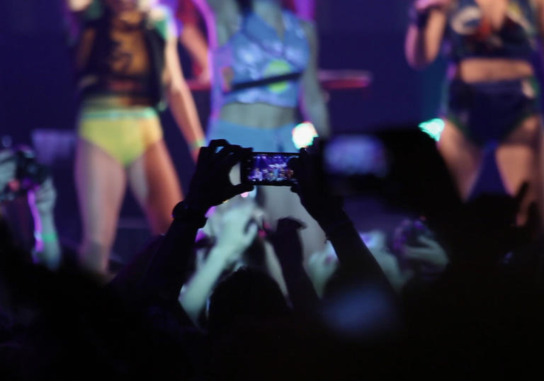 a person in a crowd holds their phone up to film the girl band on stage