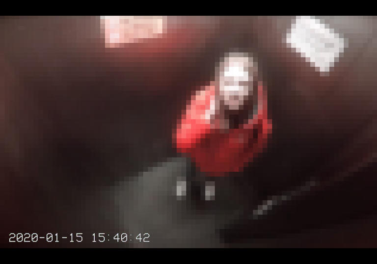A pixelated view from a CCTV camera of someone looking up at the camera in a small room. They are wearing a red coat and they have brown hair.
