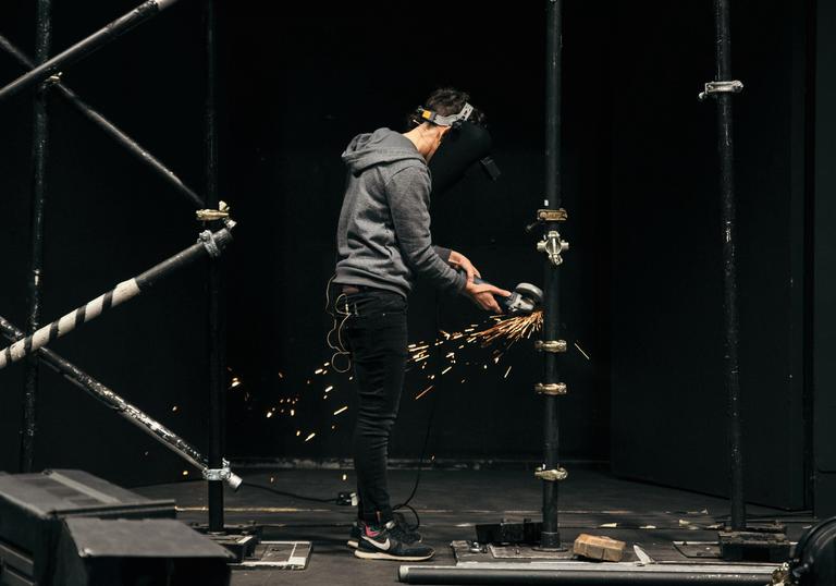 A person in a grey jumper is using a power tool on metal staging as sparks fly around them.