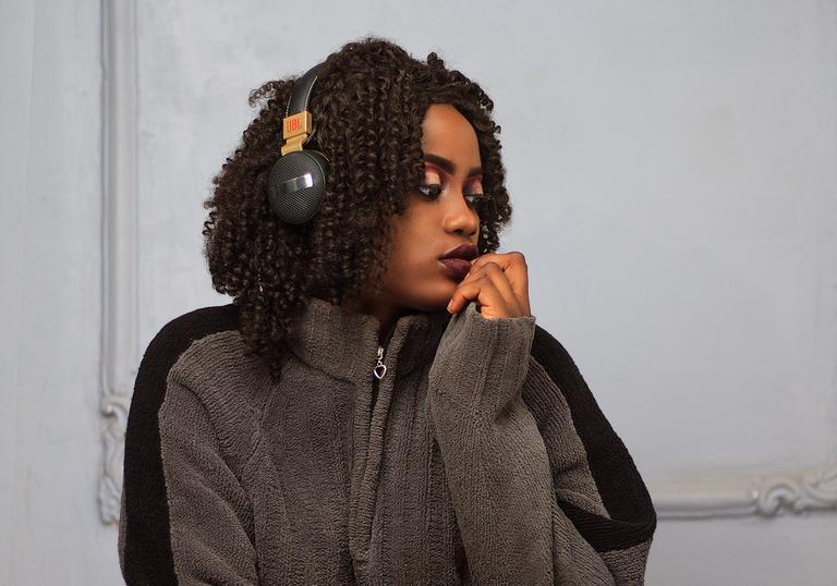 A young black woman with brown curly hair sits on the floor with headphones on looking into the distance. She's wearing a grey jumper, black tights and white trainers.