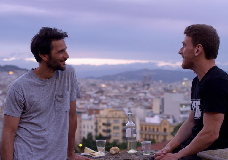 Two men gaze at each other with the violet-tinted cityscape of Barcelona as their backdrop