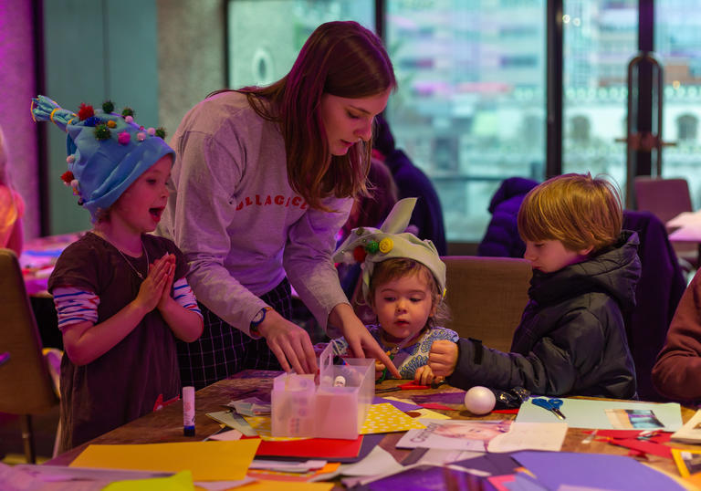 A mum with three kids, standing round a table making hats and cutting paper