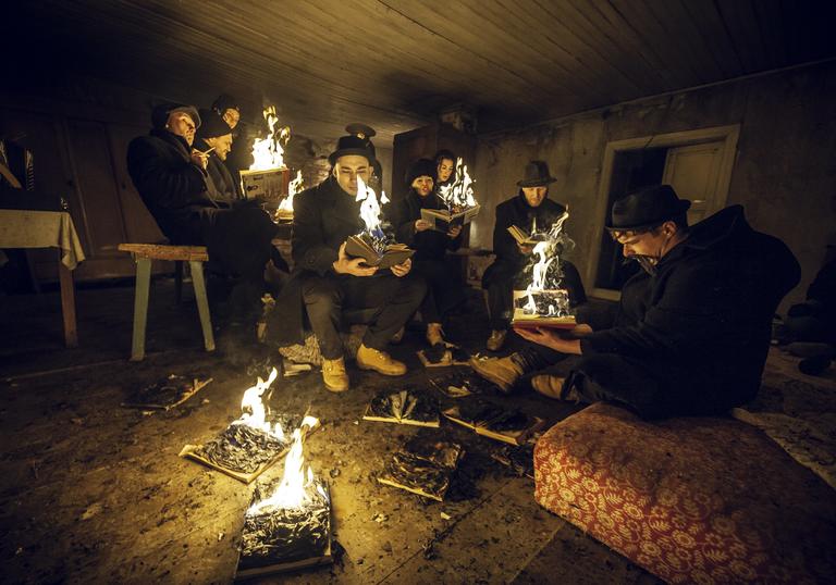 A group of people sit around in a dark room reading books that have flames emerging from the pages.