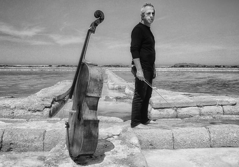 A black and white image of a pensive Giovanni standing barefoot on some ruins in a dry and barren wasteland, his cello standing upright close by