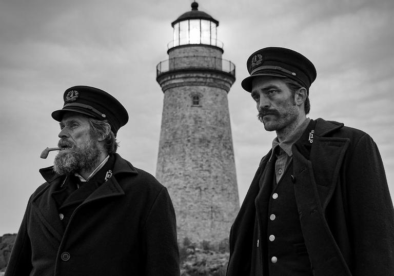 A black and white photo of Willem Dafoe and Robert Pattinson looking into the distance with a lighthouse behind them