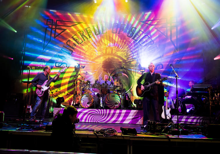 A band are on-stage at a concert with a multicoloured psychedelic backdrop.
