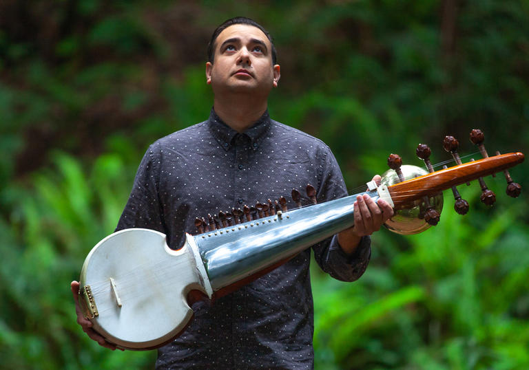 Alam Khan holding his Sarod instrument, surrounded by greenery, looking upwards