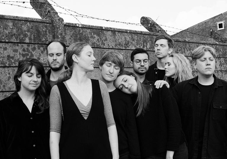 A group photo of Shards, all wearing black, with their eyes closed, standing in front of a wall with barbed wire