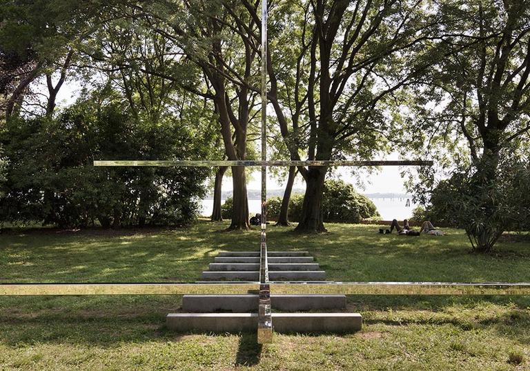 A big cross structure made from a reflective mirror like material in a nice green park by architect Carla Juaçaba